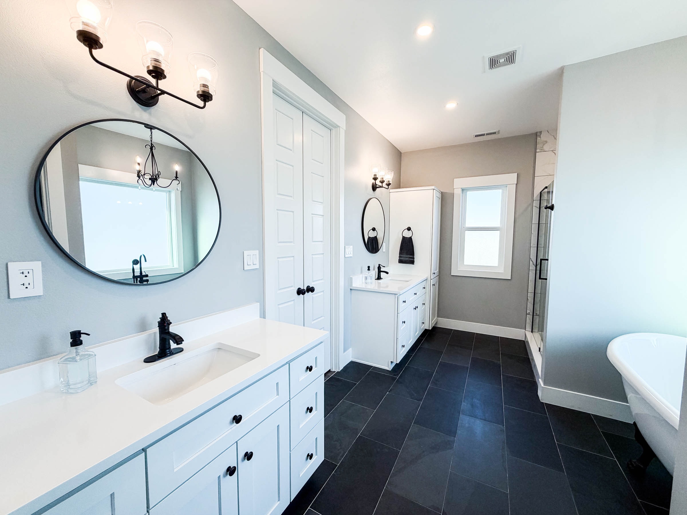 Primary bathroom dual vanities with white cabinets, black fixtures, and dark tile floors